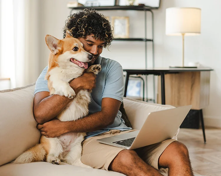 A young man hugs his corgi while they sit on a couch together and looks at a computer