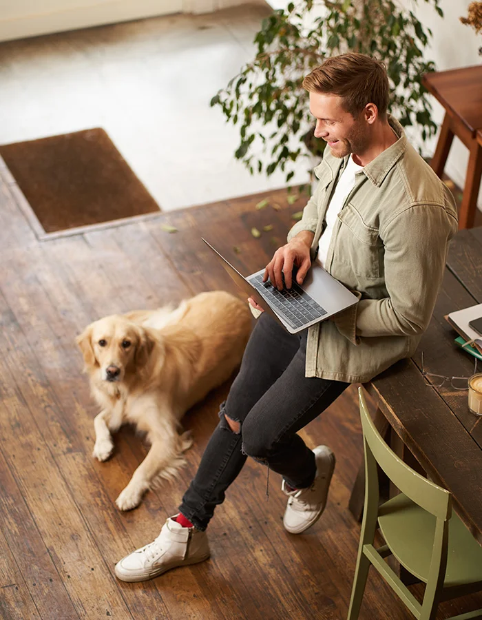 man-holding-computer-with-dog-nearby A young man stands with a laptop in his hands while his dog lays on the floor
