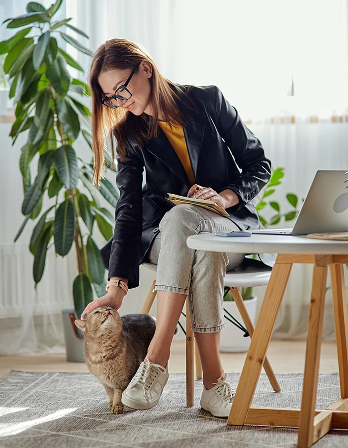 A woman leans over while sitting at her home desk to pet her cat