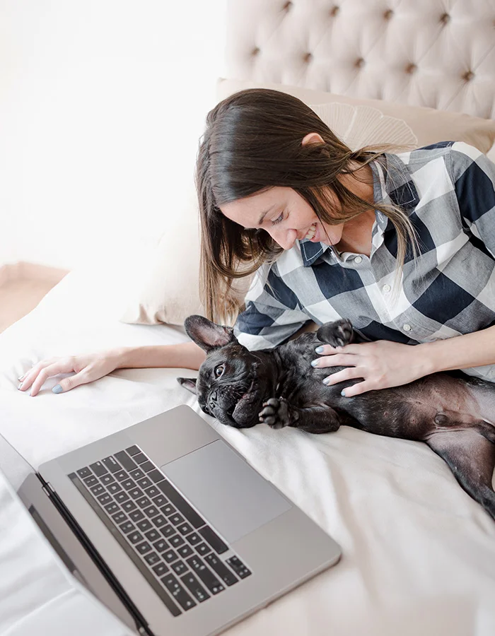 A happy woman cuddles with her french bulldog while laying on a bed in front of her computer